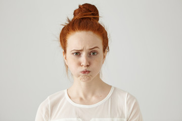 Headshot of attractive funny young female with ginger hair dressed in white blouse feeling displeased or uncomfortable with something, blowing cheeks and frowning. Human face expressions and emotions - obrazy, fototapety, plakaty