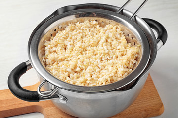 Colander with brown rice in saucepan on wooden board