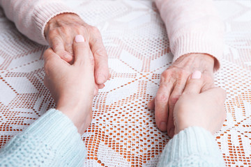 Senior woman with her caregiver at home. Holding hands, horizontally top view closeup.