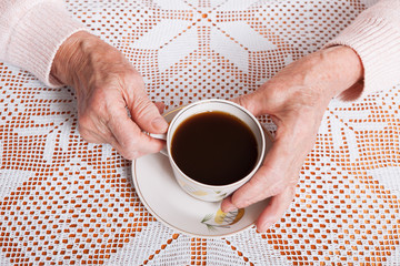 An elderly woman drinks tea at home. Senior woman holding cup of tea in their hands at table closeup. Horizontally top view
