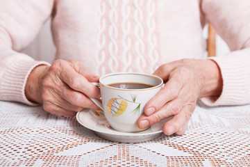 An elderly woman drinks tea at home. Senior woman holding cup of tea in their hands at table closeup