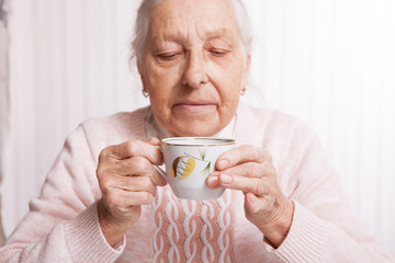 An elderly woman drinks tea at home. Senior woman holding cup of tea in their hands at table closeup