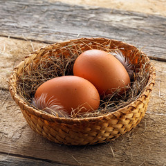 Two fresh farm eggs in a basket with hey and feather on a wooden table. Close-up shot.