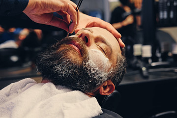 Barber shaving bearded male with a sharp razor.