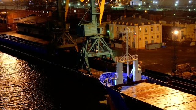 top view of empty long barge and ship on the dark water in the seaport with buildings and constructions under the lights during night