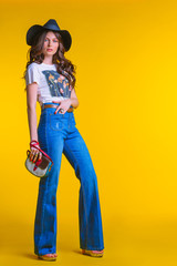 Beautiful young brunette girl in white t-shirt jeans flared with a small bag and a hat standing on a yellow background in the Studio.Fashion looks concept hats clothing elegance.