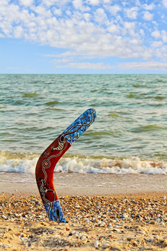Multicolored Australian Boomerang on sandy beach near sea surf and sky.