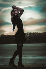 Portrait of a beautiful girl with black wings in the form of a black angel standing on the river bank that is covered with ice and sky with clouds