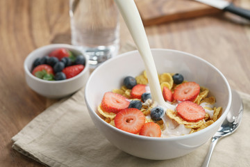 poring milk into corn flakes with berries in bowl on table