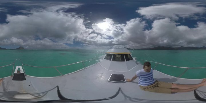 360 VR video. Man sitting on the front deck of the yacht sailing towards Mauritius on high speed. View to the ocean, captains bridge and mountains on the island