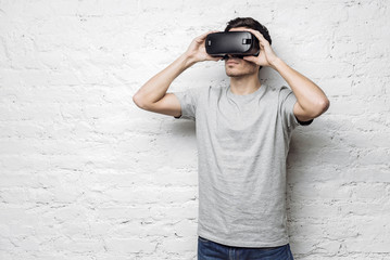 Calm and confident handsome caucasian man in casual gray t-shirt holding hands on goggles, using oculus rift headset, experiencing virtual reality while standing against white brick wall. VR concept