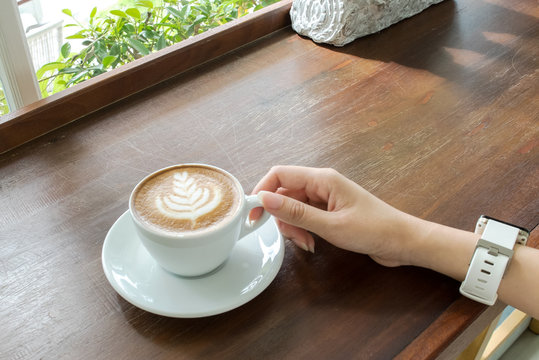 Start The Day With Good Cup Of Coffee Concept. Perspective View Business Asian Woman Hand Holding Cup Of Cappuccino Coffee With Tree Design Foam On Wooden Panel Table By Window Of Nature Garden View