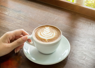 Start The Day with Good Cup of Coffee Concept. Perspective View Business Asian Woman Hand Holding Cup of Cappuccino Coffee with Tree Design Foam on Wooden Panel Table by Window of Nature Garden View