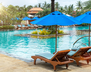 Relax by The Pool in Luxury Hotel on The Island of Thailand. Couple of Wooden Chair under The Umbrella by The View of Beautiful Luxury Swimming Pool with Various Type of Palm Trees in The Garden.