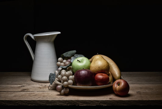 still life of various kinds of fruit and jug of water, with pictorial light on rustic wooden tables on a black background