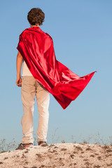 Boy playing superheroes on the sky background, teenage superhero in a red cloak on hill