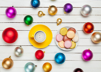photo of yellow cup of coffee and plate full of macaroons near colorful beautiful baubles on the wonderful white wooden background