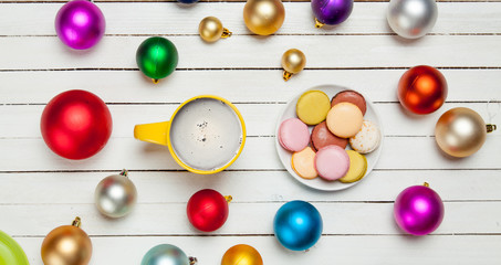 photo of yellow cup of coffee and plate full of macaroons near colorful beautiful baubles on the wonderful white wooden background