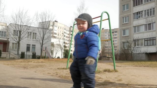 An attractive boy has been walking with his mother for 2 years in the playground in early spring. Dressed in a blue jacket and hat.