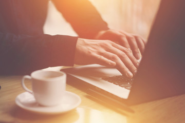 Man in black shirt working with laptop at sunny office.Modern notebook, cup of black coffee and smartphone on the wooden table.Reflections on glass surface.Horizontal, sunlight effect.
