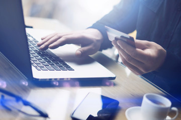 Businessman holding plastic credit card in hand and using laptop computer while sitting at the wooden table.Man making online shopping.Blurred background.Horizontal,flares.