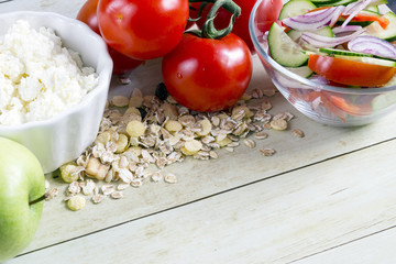Foods for healthy eating: cottage chesse, tomatoes, apple, cereal, salad on light wood background