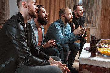 A company of four men with a beard sitting on the couch at home with beer and chips, two of them with joysticks in hand playing computer video games, the other carefully watching the game. 