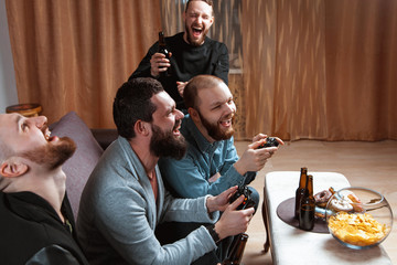 A company of four men with a beard sitting on the couch at home with beer and chips, two of them with joysticks in hand playing computer video games, the other carefully watching the game. 