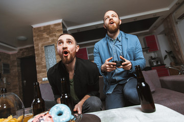 Two men with a beard sitting on the couch at home with beer and joysticks in the hands of playing computer video games,. The concept of friendship, technology and weekend