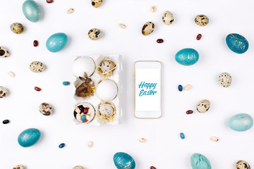 Easter eggs, candy and bunny in paper tray with smartphone on white background with blue easter eggs. Flat lay, top view