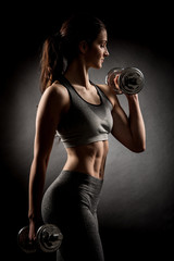 Atractive fit woman works out with dumbbells as a fitness conceptual over dark background - obrazy, fototapety, plakaty