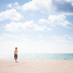 Adorable kid boy standing on the sandy beach by the ocean and looking on the gorgeous summer view. Miami summer vacations. Blue sky with clouds as a background. Child on the amazing beach.