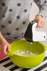 Young woman in long sleeve shirt with stars mixing and tasting dough for homemade muffins. Cooking and baking at home. Bright kitchenware and white mixer. Indoors.