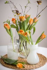 Fresh spring bunch of orange tulips and green leaves and small birds in nice cristal glass vases on the straw board and table with a tablecloth.