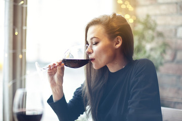 Beautiful young woman drinking red wine with friends in cafe, portrait with wine glass near window. Vocation holidays evening concept