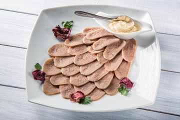 Boiled beef tongue with horseradish and herbs on white serving plate and wooden table. Selective focus