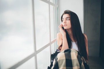 A young woman with black hair in a white T-shirt at a window flooded with sunlight uses a smartphone, communicates and searches for information on the Internet or social networks