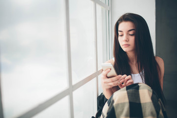 A young woman with black hair in a white T-shirt at a window flooded with sunlight uses a smartphone, communicates and searches for information on the Internet or social networks