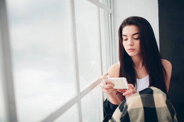 A young woman with black hair in a white T-shirt at a window flooded with sunlight uses a smartphone, communicates and searches for information on the Internet or social networks