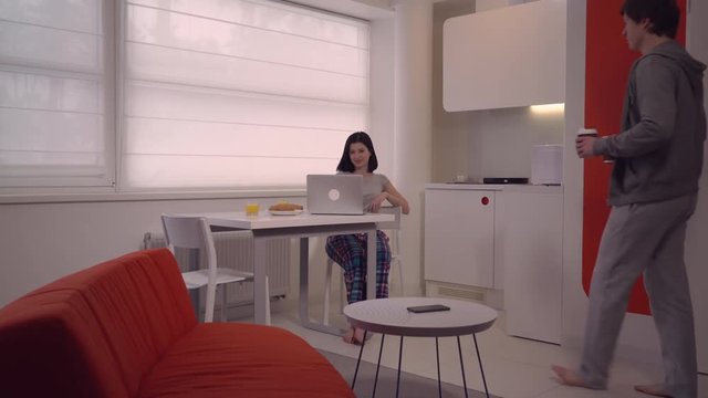 Woman sitting at the table watch funny video using computer. Man bringing coffee to go. Happy morning in weekend at home.