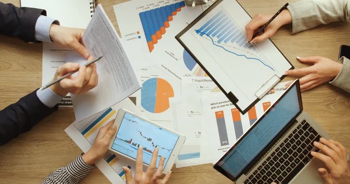 Business team are checking financial charts and graphs, pointing and working with tablet and laptop, writing notes at office wooden desk. Hands top view. Slow motion.