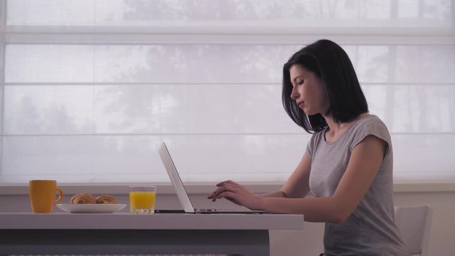 Lady working with pc at home. Caucasian busy female with black hair wearing in grey t-shirt sitting at the table with breakfast and orange juice in front of big window.