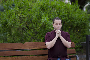 Man praying with clasped hands on a bench outside.