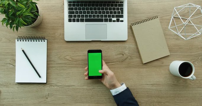 Businessman using his smartphone with green screen, scrolling news, checking financial reports, tapping on screen. Hands top view. Office desk background. Slow motion. red epic