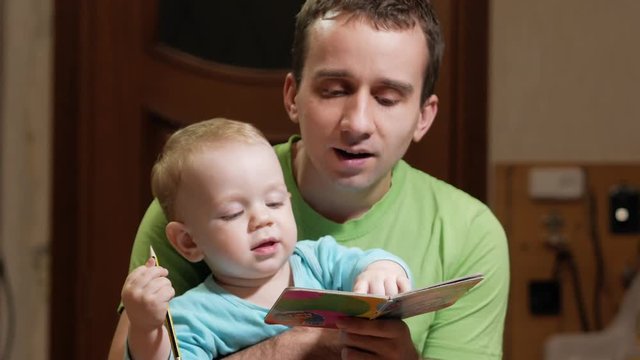 An attractive boy 2 years old and father read a book at home. Dad shows the pictures and the son calls the animals.