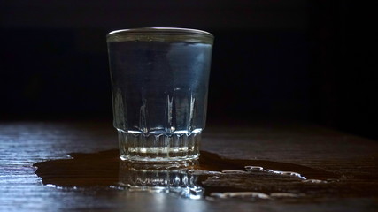 A glass of vodka on wooden table. Part of vodka poured off side. Dark background
