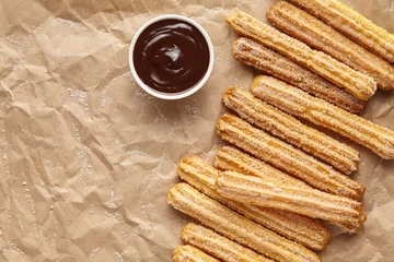 Schilderijen op glas Churros traditional homemade Spain breakfast or lunch meal street fast food baked sweet dough snack dessert with chocolate dipping on rustic decorative parchment paper background. Flat lay top view © GreenArt Photography