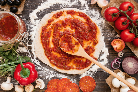 Cooking pizza. Adding fresh tomato sauce to pizza dough. Pizza ingredients on the wooden table top view
