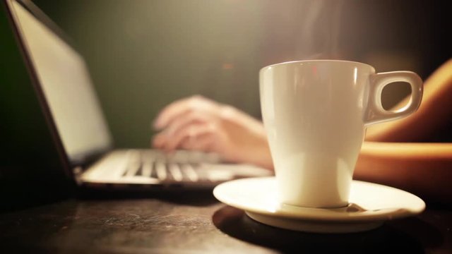 Still shot of a woman's hands typing on a laptop keyboard, and drinking a cup of hot coffee.