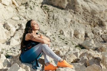 Young woman posing near the beautiful mountains dressed colorful clothes sitting on a stone